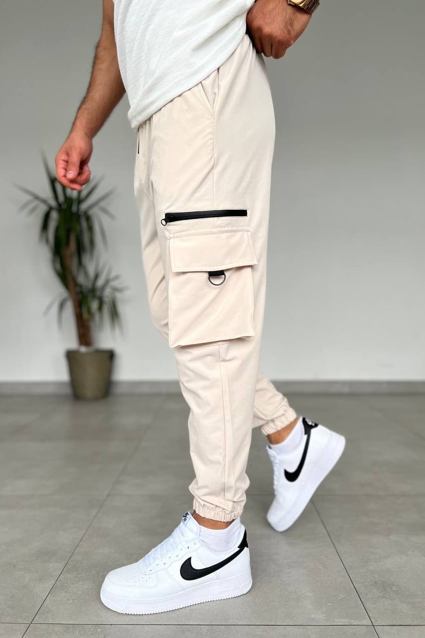 Cargo Jogger Pants - Tapered cargo jogger pants in a sand beige finish, designed with a modern streetwear silhouette and elastic cuffed ankles. Crafted from lightweight durable fabric with an adjustable waistband for all-day comfort. Features spacious side cargo pockets, zip utility detail, and functional storage for a clean urban look. Perfect with tees, hoodies, and sneakers.