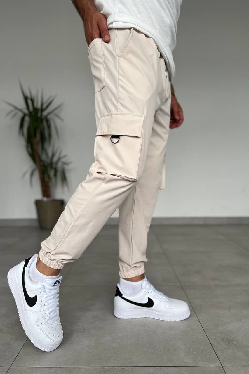 Cargo Jogger Pants - Tapered cargo jogger pants in a sand beige finish, designed with a modern streetwear silhouette and elastic cuffed ankles. Crafted from lightweight durable fabric with an adjustable waistband for all-day comfort. Features spacious side cargo pockets, zip utility detail, and functional storage for a clean urban look. Perfect with tees, hoodies, and sneakers.
