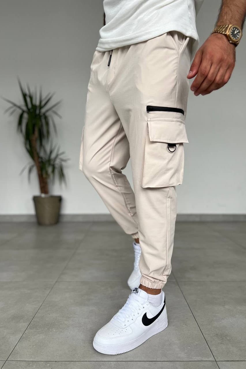 Cargo Jogger Pants - Tapered cargo jogger pants in a sand beige finish, designed with a modern streetwear silhouette and elastic cuffed ankles. Crafted from lightweight durable fabric with an adjustable waistband for all-day comfort. Features spacious side cargo pockets, zip utility detail, and functional storage for a clean urban look. Perfect with tees, hoodies, and sneakers.